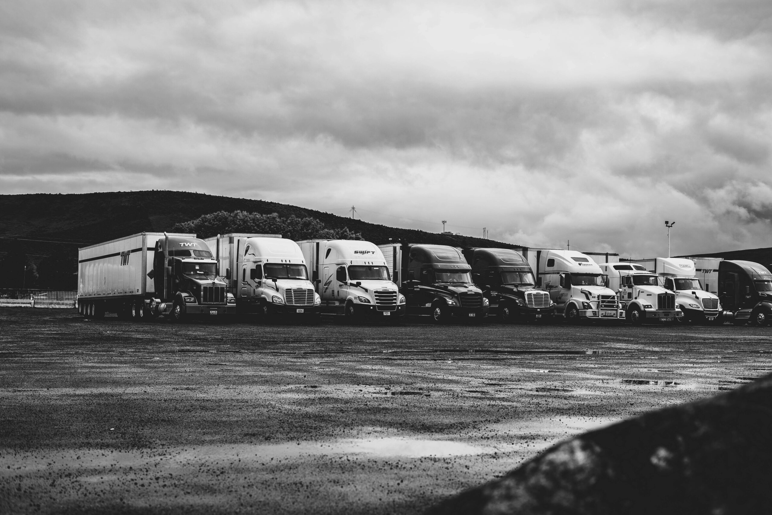 pexels-photo-2348359-2348359-1 Row of parked semi trucks in a rainy lot, captured in a dramatic black and white setting.