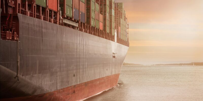 A colossal cargo ship loaded with containers navigates through calm waters against a vibrant sunset sky.