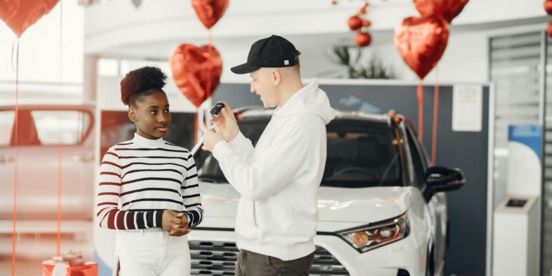 A couple exchanging car keys inside a showroom decorated with heart-shaped balloons, celebrating a new car purchase.