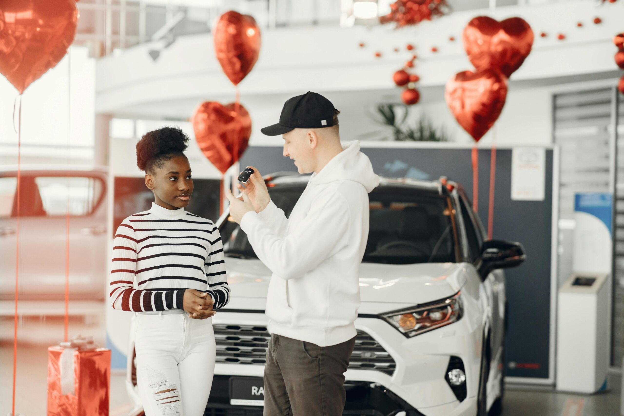 A couple exchanging car keys inside a showroom decorated with heart-shaped balloons, celebrating a new car purchase.