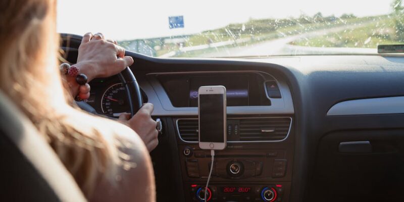 View from behind a woman driving a car on a sunny day with a phone mounted on the dashboard.